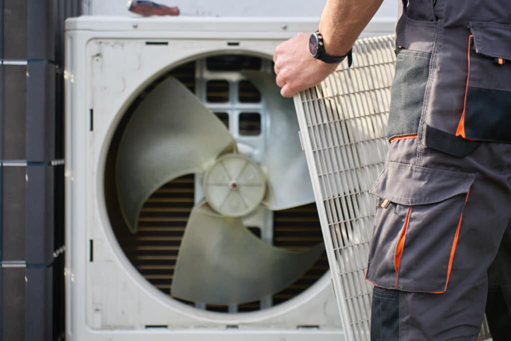 A technician in gray work pants with orange accents is servicing an outdoor air conditioning unit. He is holding a protective cover with a fan visible in the background, highlighting the appliance's mechanical components.