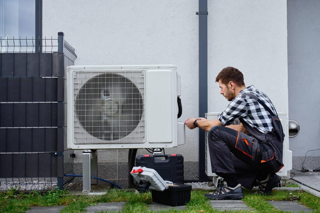 A technician in a checked shirt and overalls works on an air conditioning unit outside a building. Tools are placed next to him on the grass, and a fence is visible in the background.