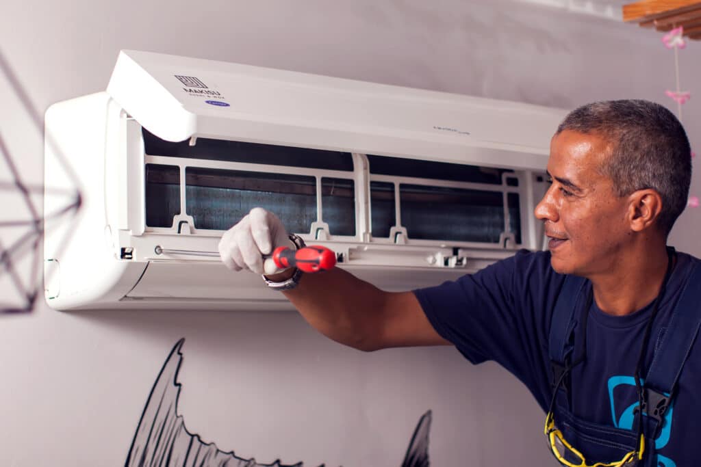 A technician wearing gloves is working on an air conditioning unit mounted on a wall. He is using a screwdriver to access the unit, which is white and has a modern design. The background features a stylized wall drawing.