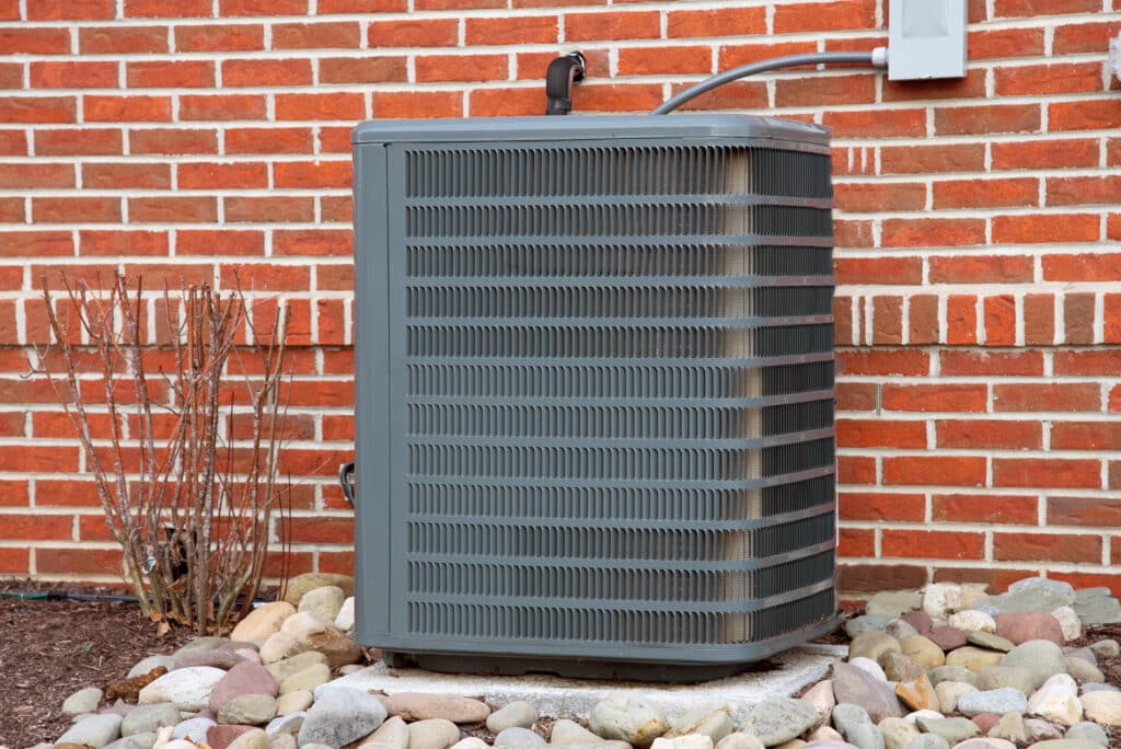 An outdoor air conditioning unit situated next to a brick wall. The unit is gray with a grid pattern on its sides. Surrounding the unit are small rocks and a bare shrub. A power line is connected to the unit, extending towards the wall.