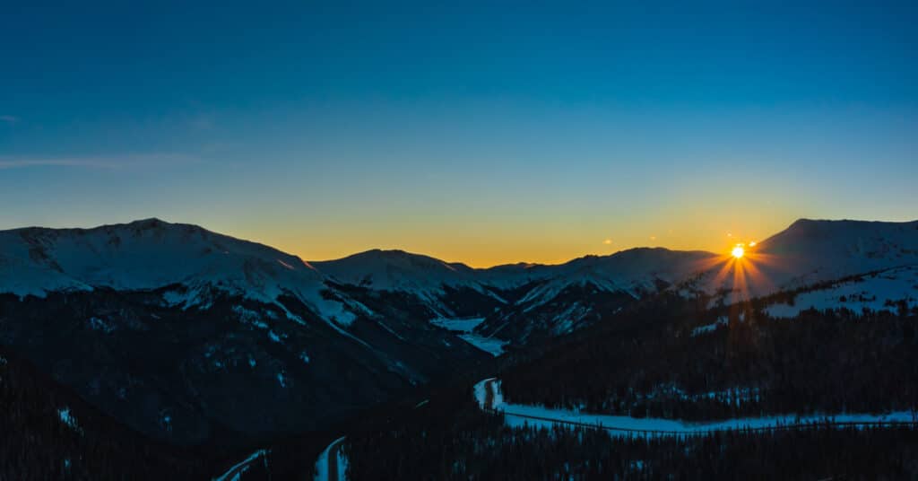 A panoramic view of snow-covered mountains at sunrise. The sky transitions from deep blue to warm orange near the horizon, with the sun peeking out from behind the peaks. The landscape features valleys and a winding river in the foreground.