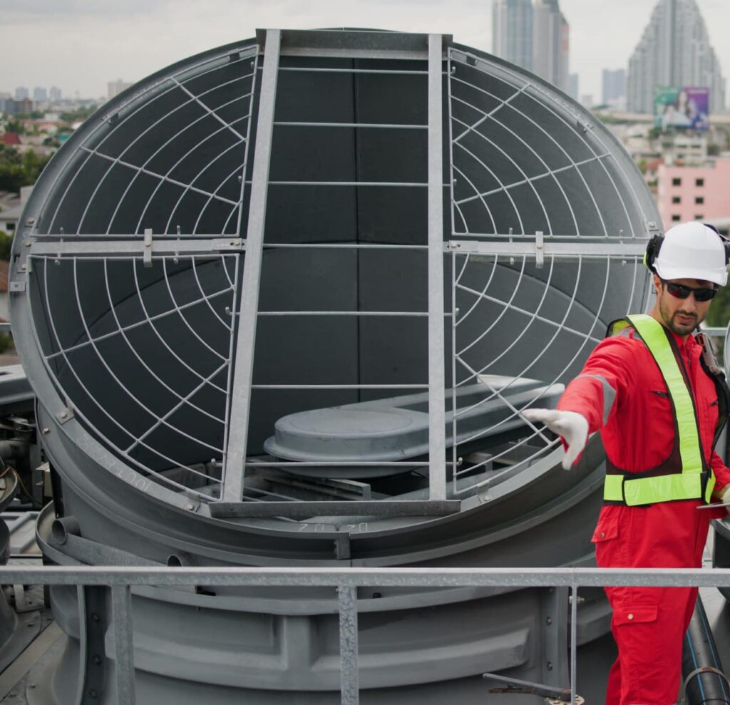 A technician wearing a red jumpsuit and helmet stands near a large, circular structure with a mesh cover on top. He is gesturing with his right hand, and a cityscape is visible in the background under a cloudy sky.