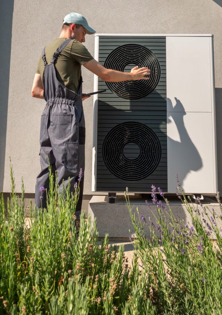 A technician in a green shirt and gray overalls is inspecting a wall-mounted heat pump unit. He is touching the upper circular component while standing in front of lavender plants and a concrete wall.