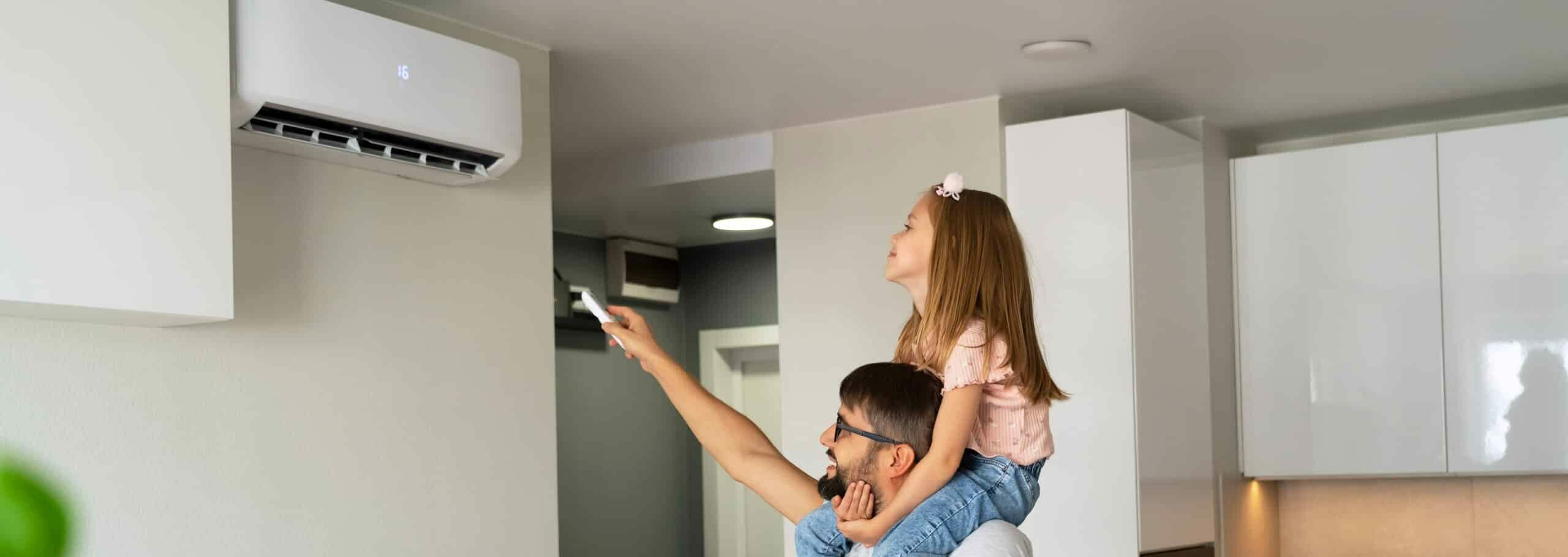 A man holding a remote control points toward a wall-mounted air conditioner while a young girl sits on his shoulders, looking up. The background features a modern kitchen setting with light-colored cabinets.
