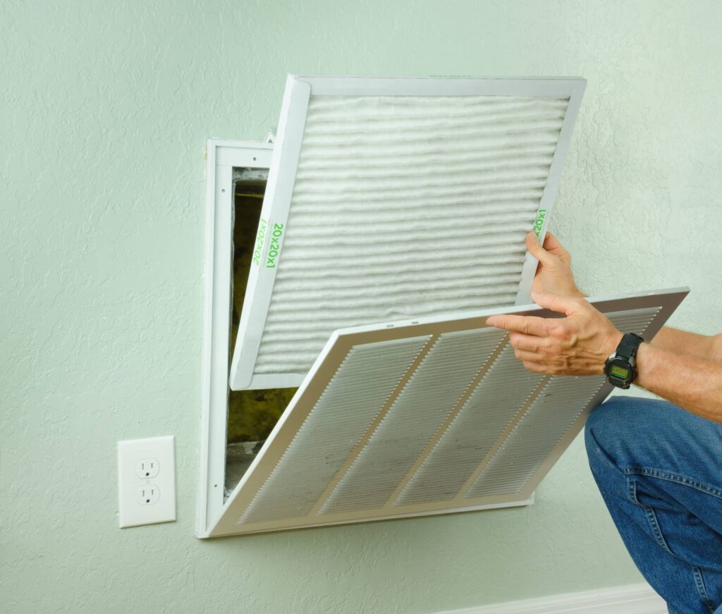 A person removing an air filter from a wall-mounted HVAC system. The filter has a white frame with pleated material, and the wall is painted a light green. An electrical outlet is visible beside the unit.