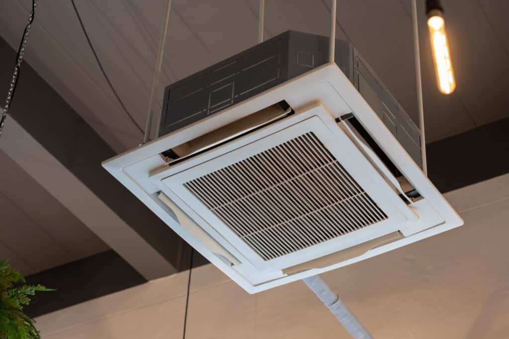 An overhead ceiling-mounted air conditioning unit with a white exterior, featuring a grid pattern for airflow. It is suspended from the ceiling in a modern interior space, with industrial-style lighting visible in the background.