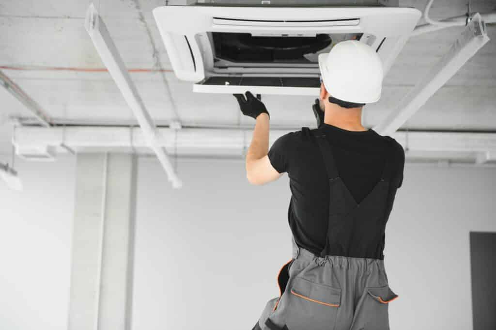 A maintenance worker wearing a hard hat and gloves is repairing an air conditioning unit mounted on the ceiling. The worker is reaching up to manage the unit, and the background is a modern ceiling with exposed ducts.