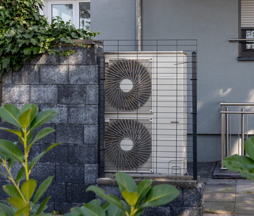 An outdoor air conditioning unit mounted against a gray wall, featuring a protective metal grate. The unit has two large fans and is surrounded by greenery, with a small hedge and plants in the foreground.