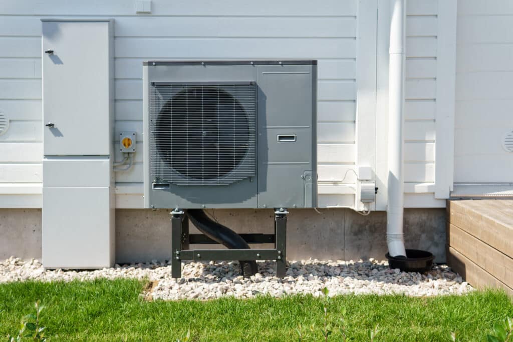 An outdoor heat pump unit placed on a stone bed next to a residential building. The unit features a large circular fan and is mounted on a metal frame. Nearby, there's an electrical box and white drain pipe against a white wall.