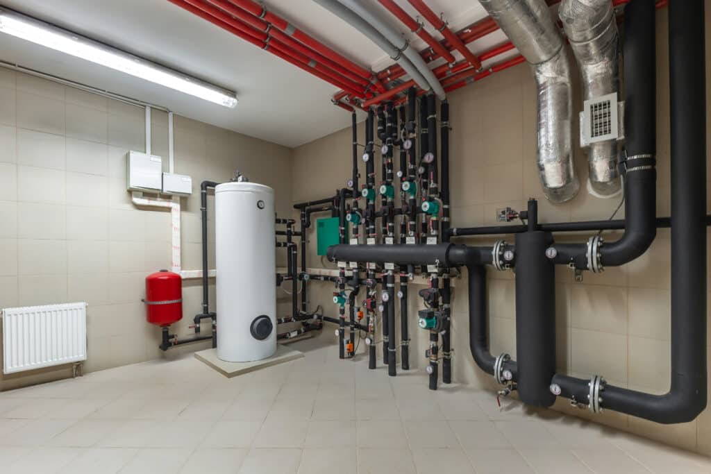 An industrial plumbing room featuring a white hot water tank, various black pipes, green valves, and red piping along the wall. A red water pressure tank is visible, along with heating equipment and a radiator mounted on a tiled floor.
