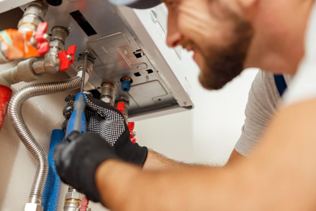 A technician wearing a grey shirt and gloves is working on a water heater. He is using a tool to adjust connections, with pipes and fittings visible in the background. Red and blue valve handles can be seen alongside the plumbing setup.