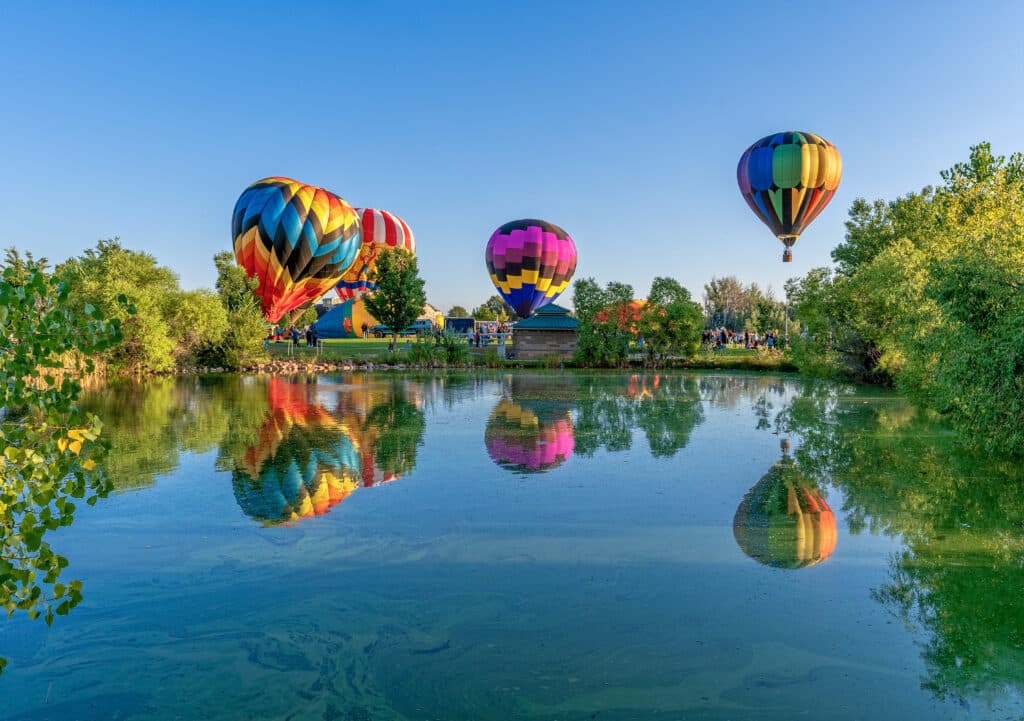 Colorful hot air balloons are inflated and floating above a calm lake surrounded by greenery. The reflections of the balloons and trees are visible on the water's surface under a clear blue sky.