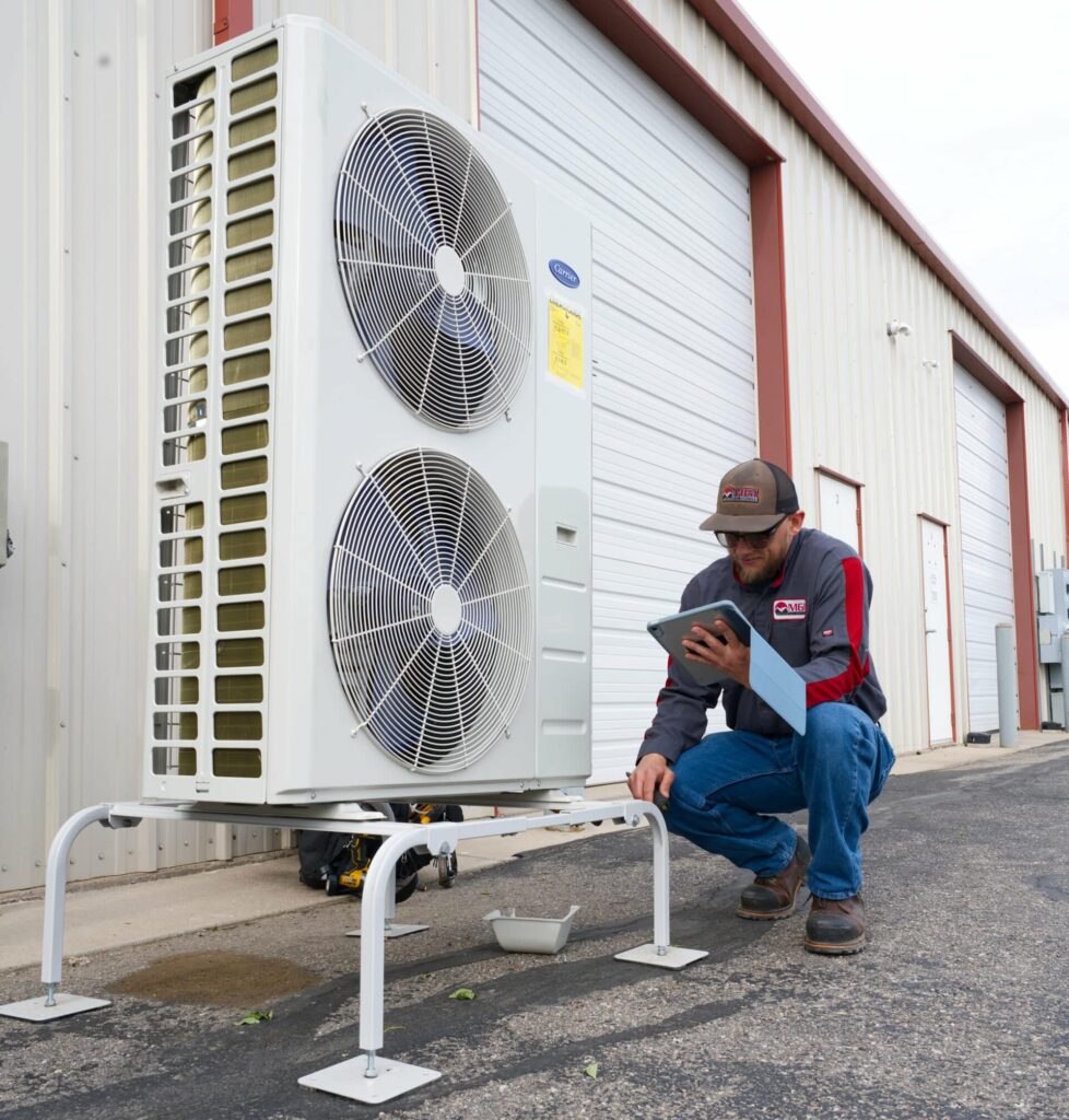 A technician in a cap kneels next to a large outdoor air conditioning unit with two fans. He is holding a tablet and examining the unit, set on a concrete surface against a backdrop of a metal building with roll-up doors.