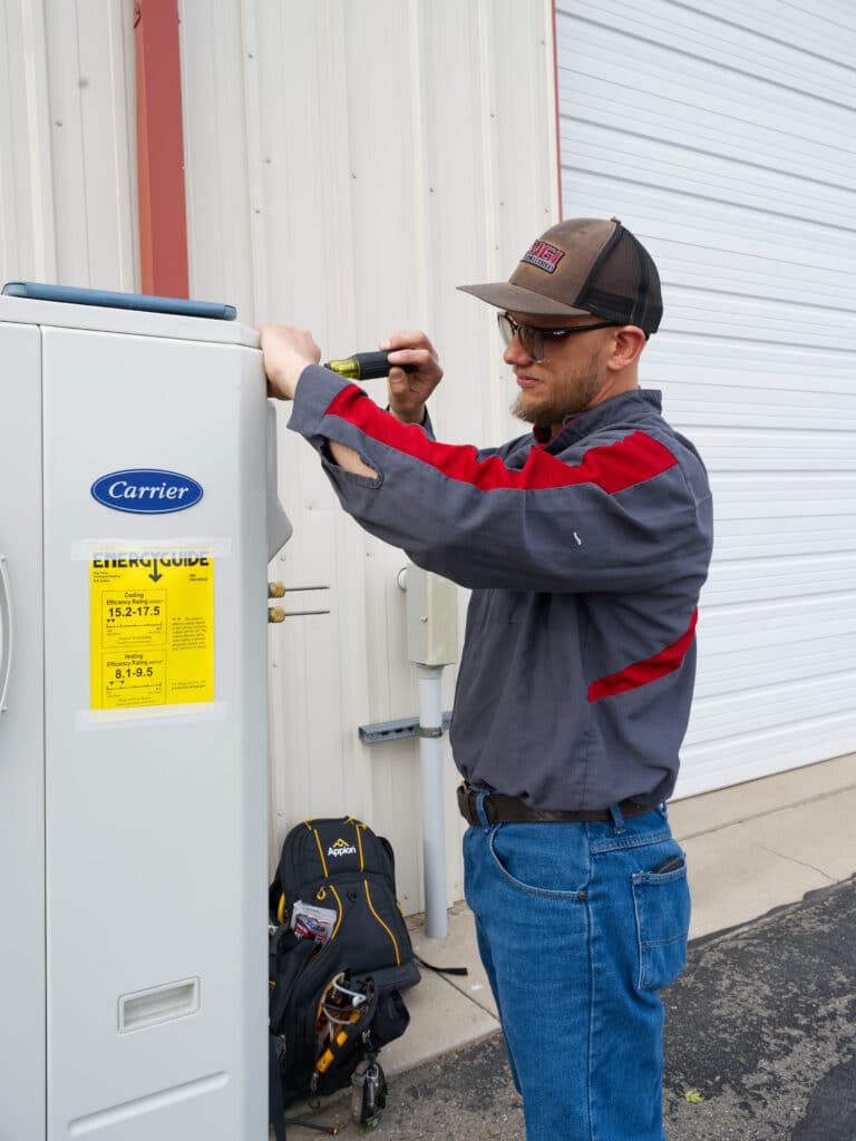 A man in a gray work shirt with red accents and a cap is using a screwdriver to work on a Carrier appliance. A black toolbox is placed on the ground beside him, and a building with a large white door is visible in the background.