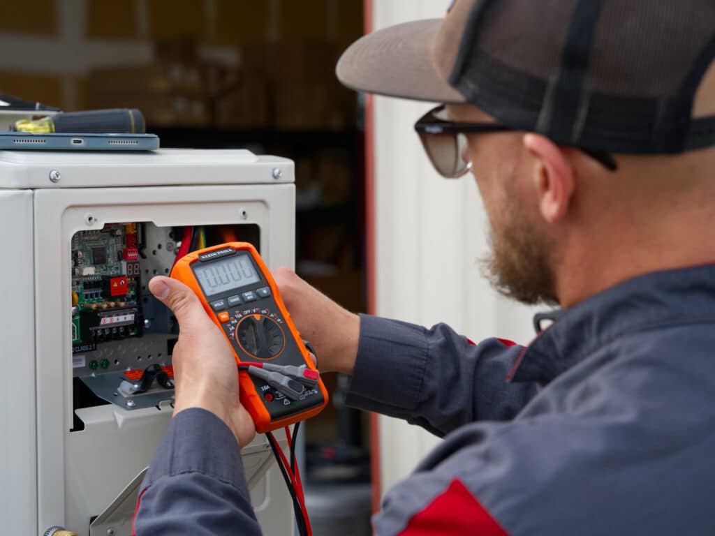 A technician wearing a baseball cap and glasses uses a multimeter to check electrical connections inside a control panel. The multimeter is orange, and the panel contains several circuit boards and wires.