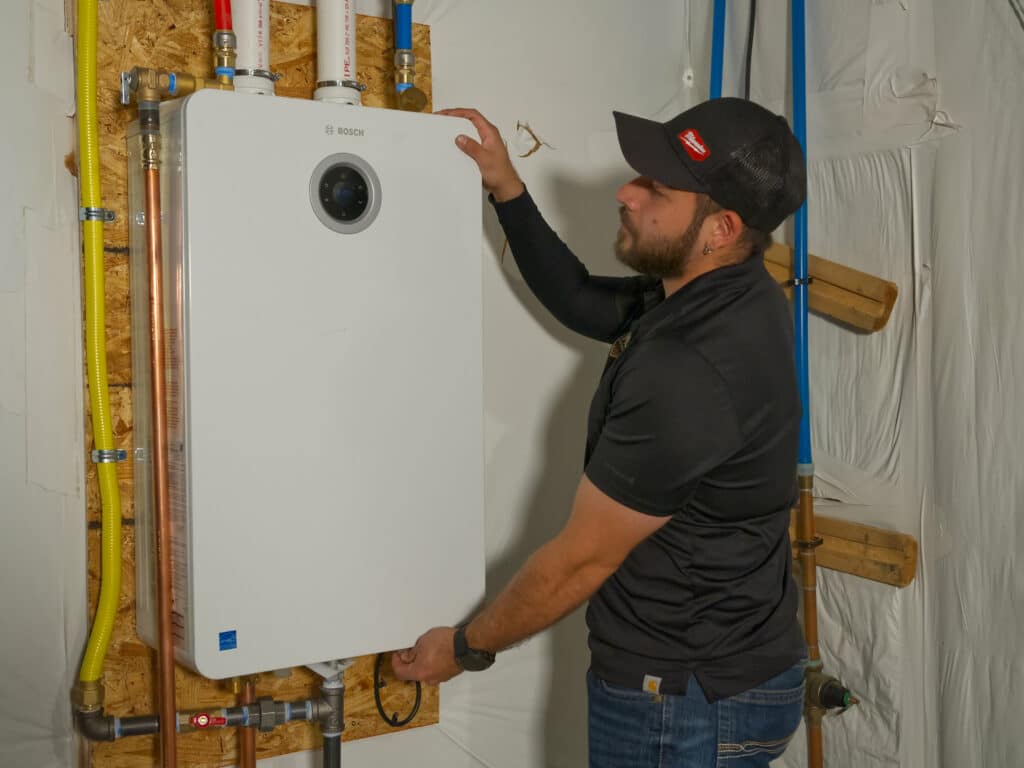 A man in a black shirt and cap is installing a white tankless water heater mounted on a wooden wall. The installation area includes pipes in various colors, including blue and yellow, and insulated walls.