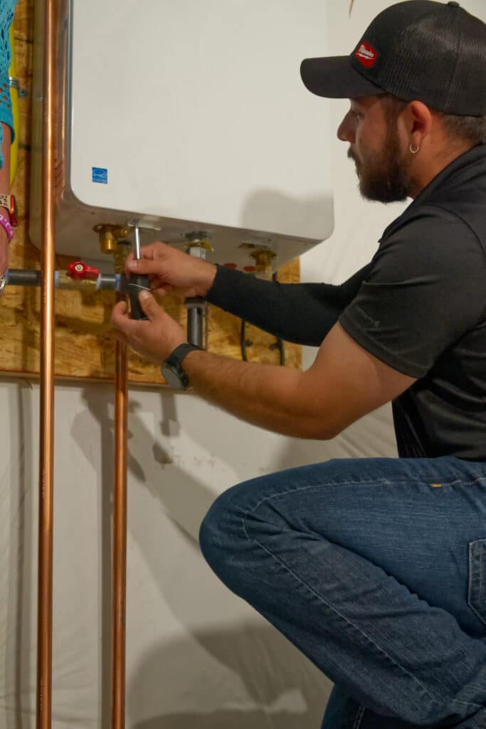 A man wearing a black cap and a dark shirt kneels while tightening a connector on copper piping. A tankless water heater is mounted on the wall behind him, with various pipes connected to it.