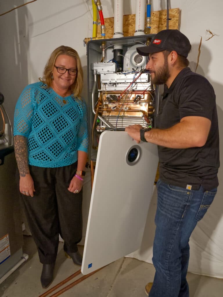 A woman with long hair and glasses stands next to a man in a black shirt and cap, who is holding a white panel near a large boiler or heating system in a basement. Colorful pipes are visible on the wall behind them.