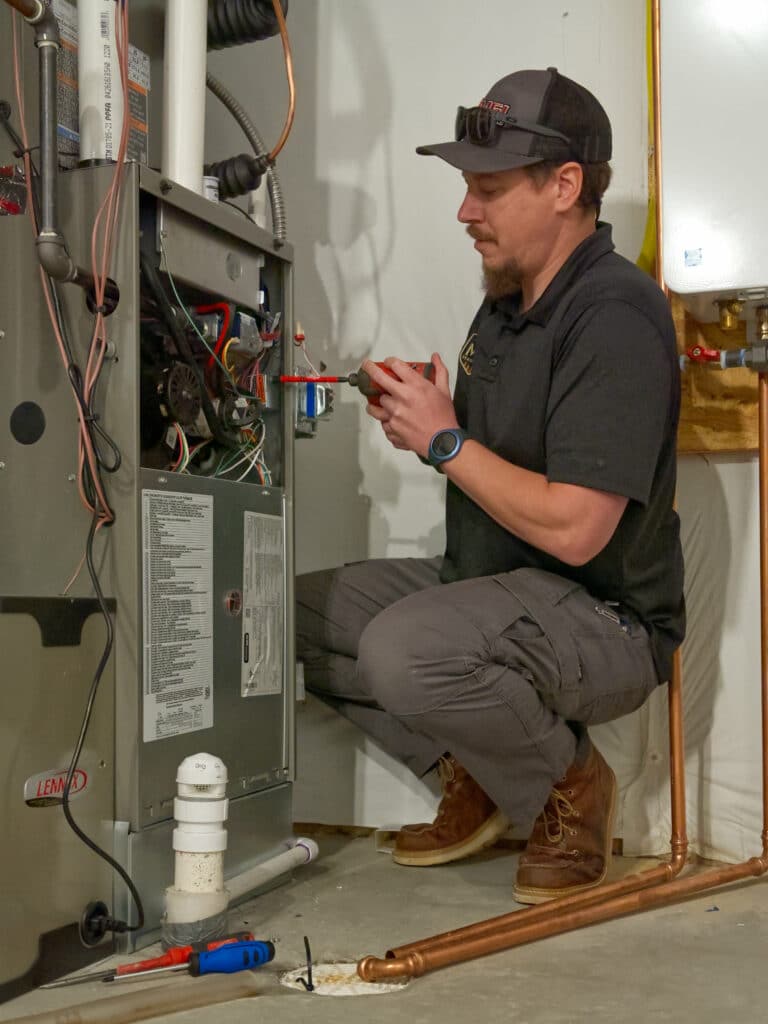 A technician in a dark shirt and baseball cap kneels beside a furnace, adjusting wires and components inside it. Tools are visible on the floor, including a screwdriver and piping nearby, with a white wall and heating equipment in the background.