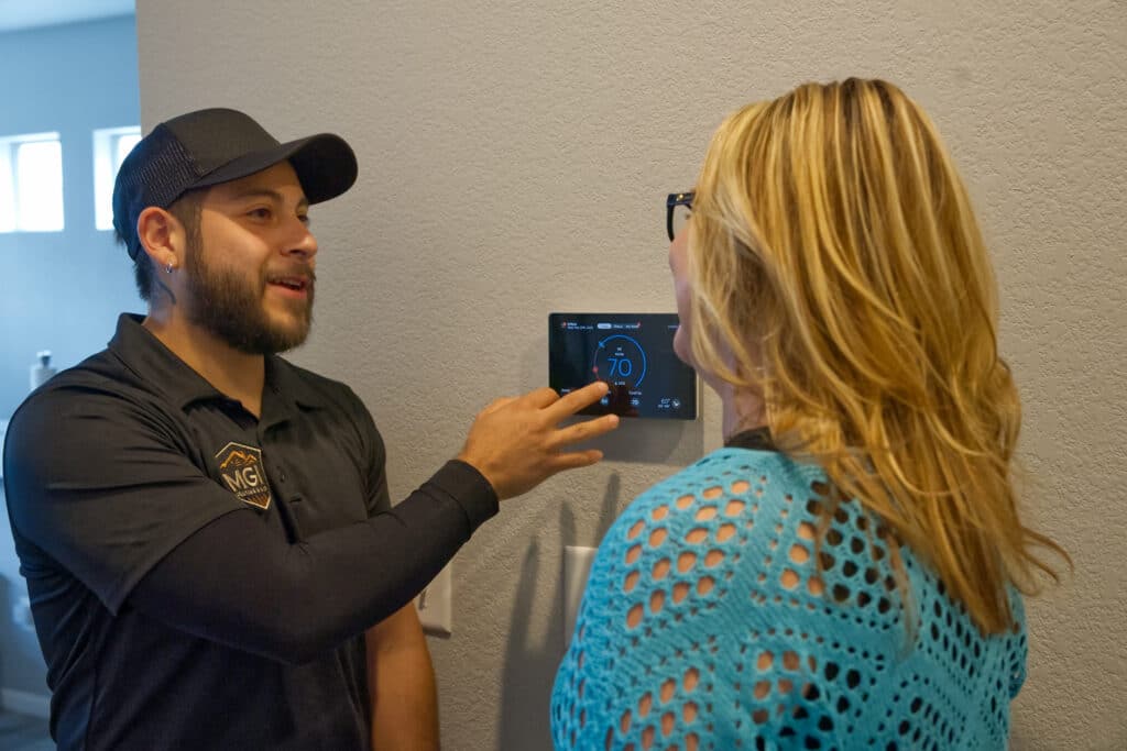 A man in a black shirt points to a digital thermostat on a wall while explaining its features to a woman with long hair wearing glasses and a blue sweater. The background includes a light-colored wall and windows letting in natural light.