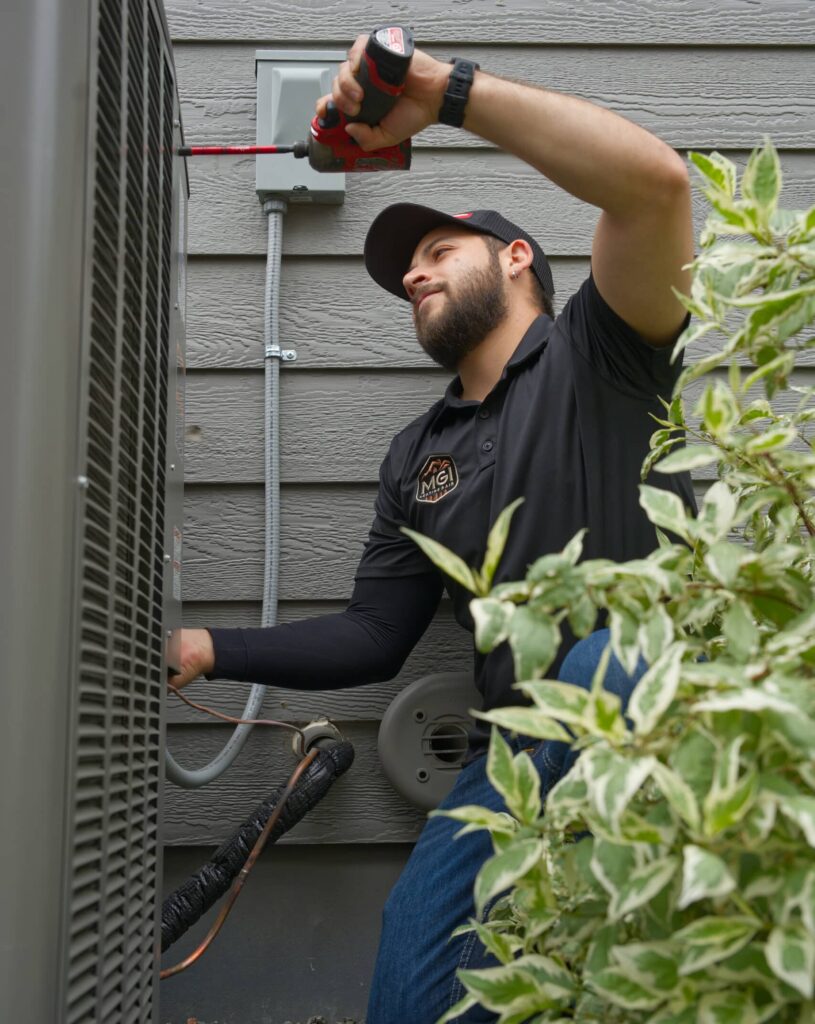 A technician wearing a black shirt and cap is using a screwdriver to work on an air conditioning unit outside. The unit is beside a wall with electrical connections, and green plants are visible in the foreground.