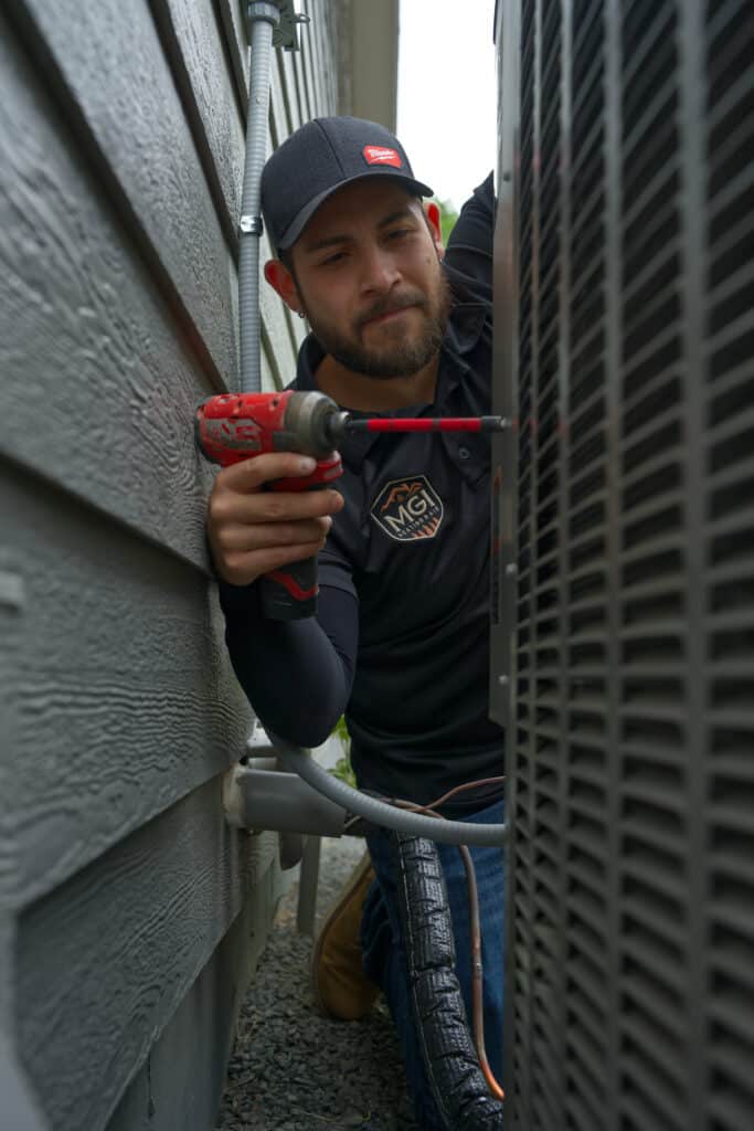 A technician using a cordless screwdriver to work on an air conditioning unit positioned against a house. The technician is dressed in a black shirt and baseball cap, focusing on the equipment in a narrow space between the building and the unit.