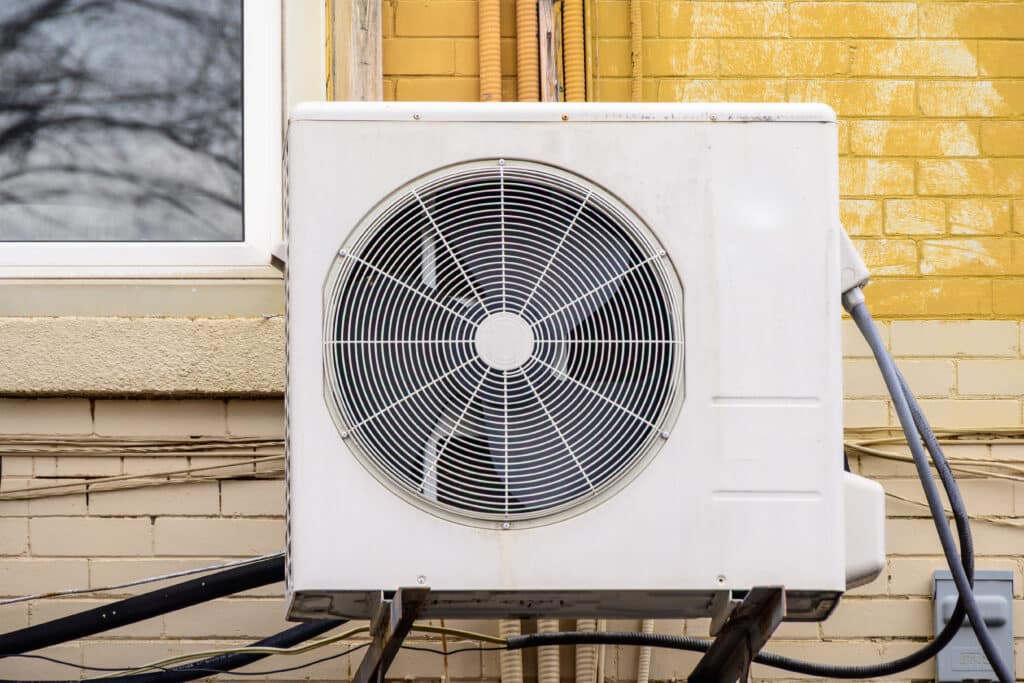 An outdoor air conditioning unit mounted on a wall near a window. The unit has a large, round metal fan covered with a circular grille, with power cables and pipes visible along the wall.
