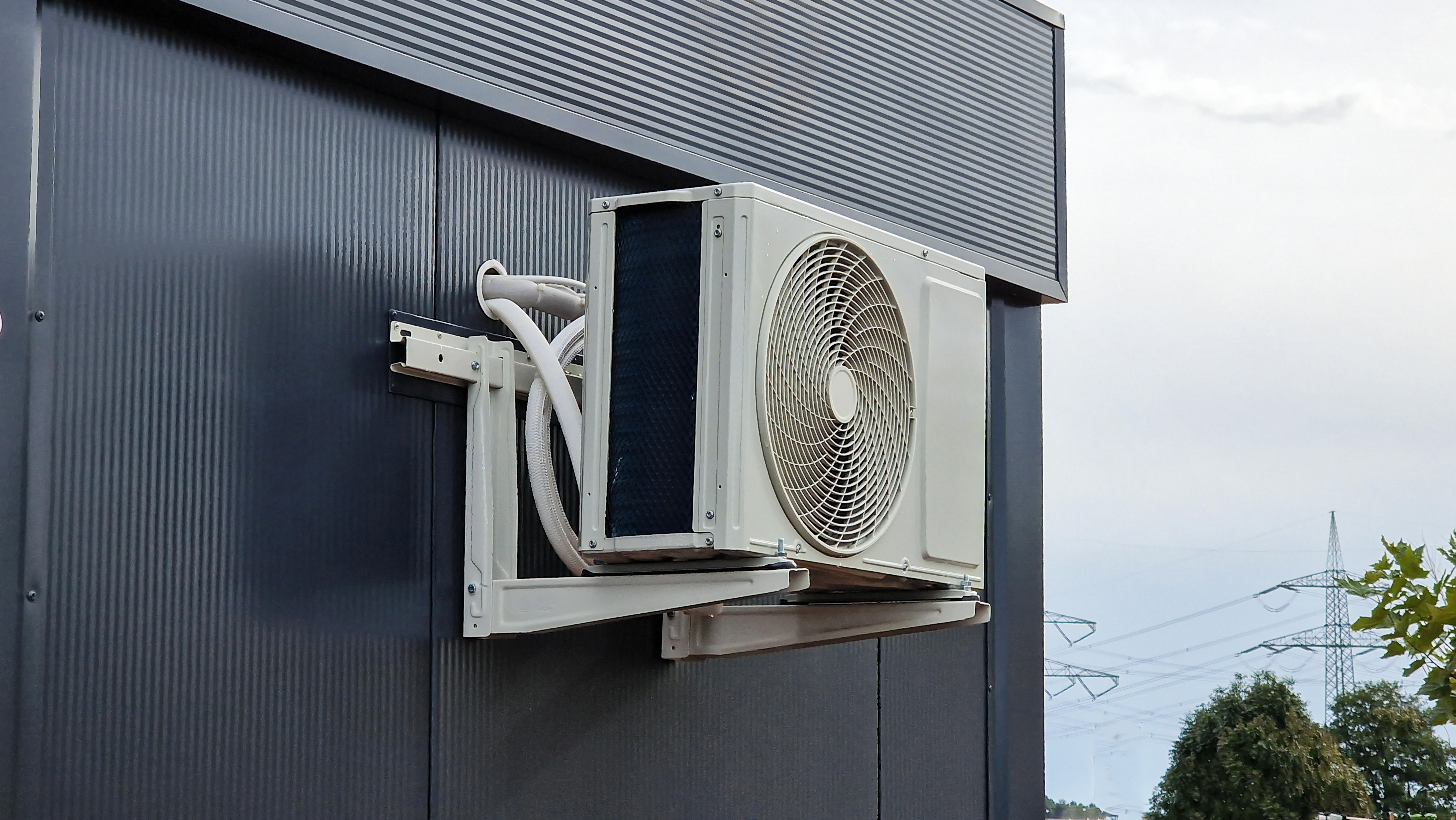 An exterior wall-mounted air conditioning unit is installed against a textured black wall. The unit has a white fan casing and is connected to pipes leading into the building. The background includes trees and power lines in the distance.