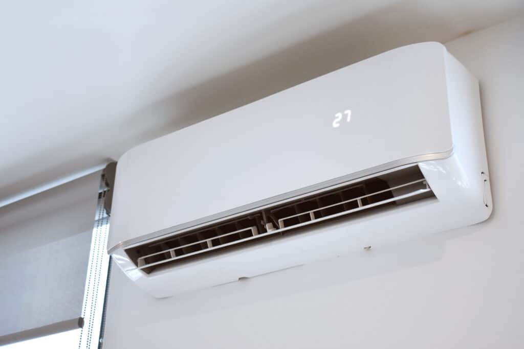 A white air conditioning unit mounted on a wall, displaying a temperature reading of 27 degrees. The unit features two ventilation slits for airflow, with a window blind partially visible in the background.