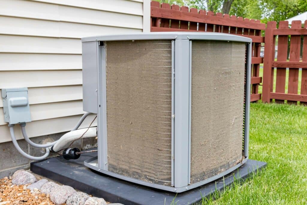 A central air conditioner unit sits on a concrete pad beside a house, with a grassy yard and red wooden fence in the background.