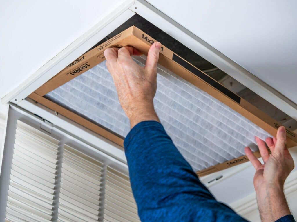 A person installs a rectangular air filter in a ceiling vent, with a louvered return grille visible below.
