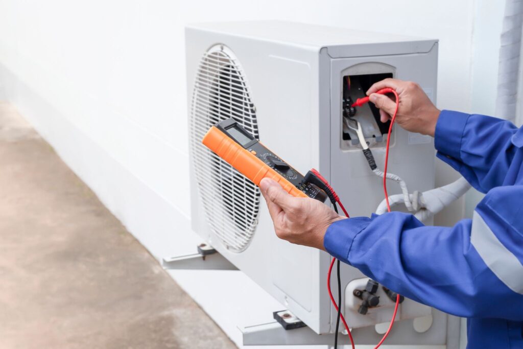 A technician in a blue uniform uses a multimeter to test wiring inside an outdoor air conditioning unit beside a white wall.