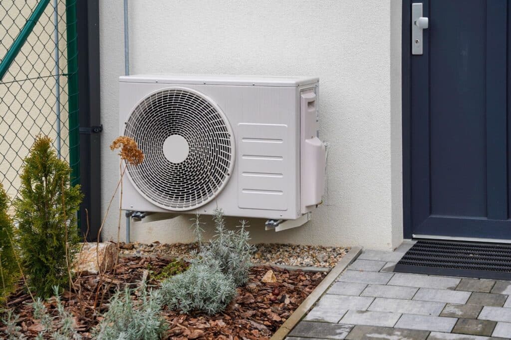 A wall-mounted outdoor air conditioner unit beside a dark blue door, with a paved walkway and small plants in front of the house.