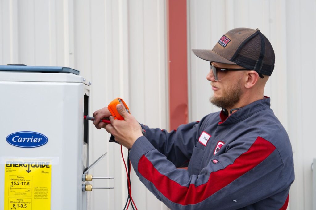 A technician in a cap and work jacket uses a multimeter to check a Carrier outdoor HVAC unit beside a corrugated metal wall.