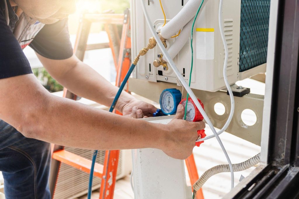 A technician adjusts pressure gauges and hoses on an outdoor HVAC unit, with orange ladders and sunlight in the background.